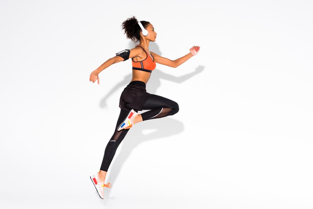 vista lateral da bela atleta afro-americana atlética em fones de ouvido e pular braçadeira smartphone em branco
 - Foto, Imagem