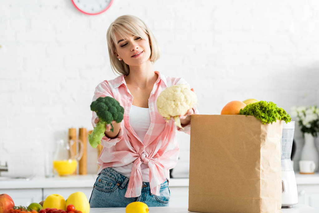 cheerful blonde girl holding cauliflower and broccoli near paper bag - Photo, Image