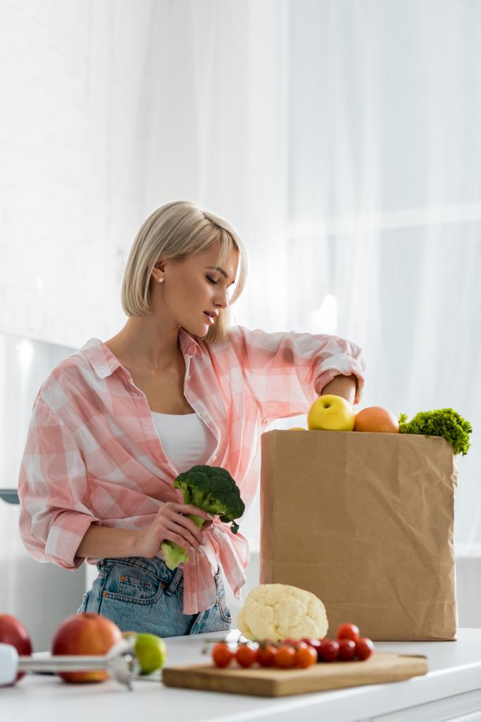 atractiva chica rubia sosteniendo brócoli orgánico cerca de bolsa de papel
 - Foto, Imagen