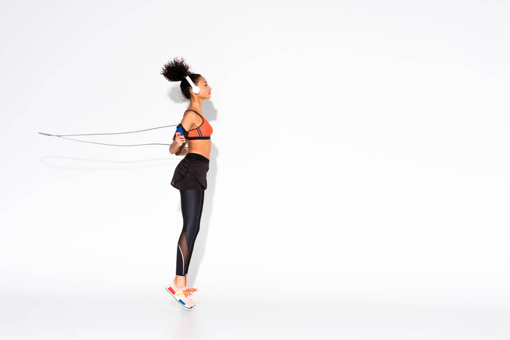 atlética afro-americana esportista em fones de ouvido exercitando-se com pular corda no branco
 - Foto, Imagem