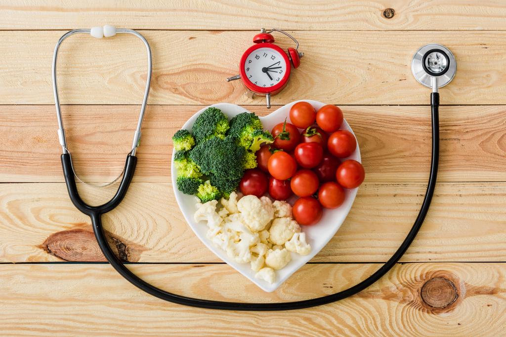 Widok z góry smaczne warzywa na talerzu kształt serca w pobliżu stetoskop i retro budzik na drewnianej powierzchni  - Zdjęcie, obraz