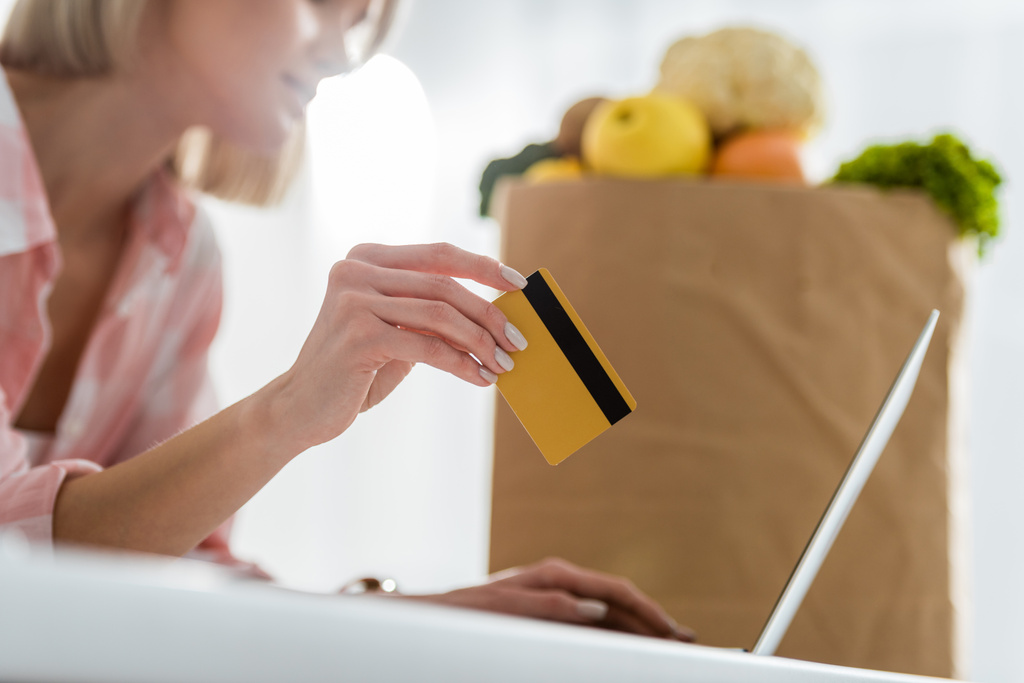 cropped view of girl holding credit card while using laptop near paper bag with groceries   - Photo, Image
