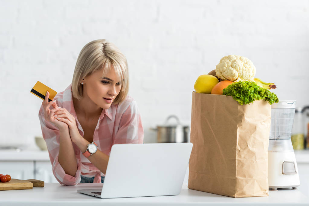 gyönyörű szőke lány Holding hitelkártya használat közben laptop közelében papírzacskót az Élelmiszeri   - Fotó, kép