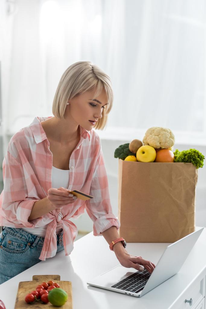 Blondine hält Kreditkarte in der Hand, während sie Laptop in der Nähe von Papiertüte mit Lebensmitteln benutzt   - Foto, Bild