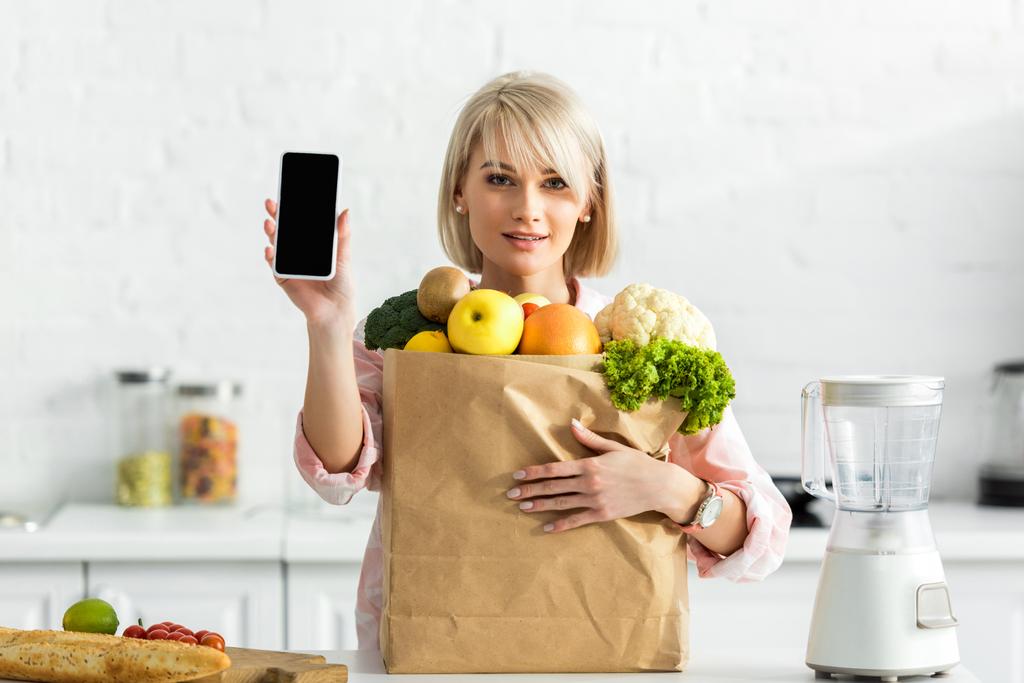 ブロンドの女の子は食料品と紙袋の近くに空白の画面でスマートフォンを保持    - 写真・画像