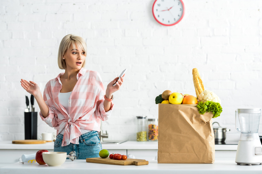 zaskoczona blond dziewczyna ze smartfonem w pobliżu torby papierowej z artykułów spożywczych  - Zdjęcie, obraz