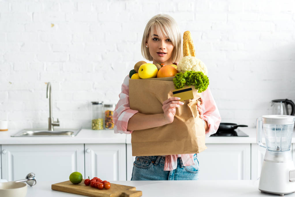 удивленная блондинка с кредиткой рядом с бумажным пакетом с продуктами
  - Фото, изображение