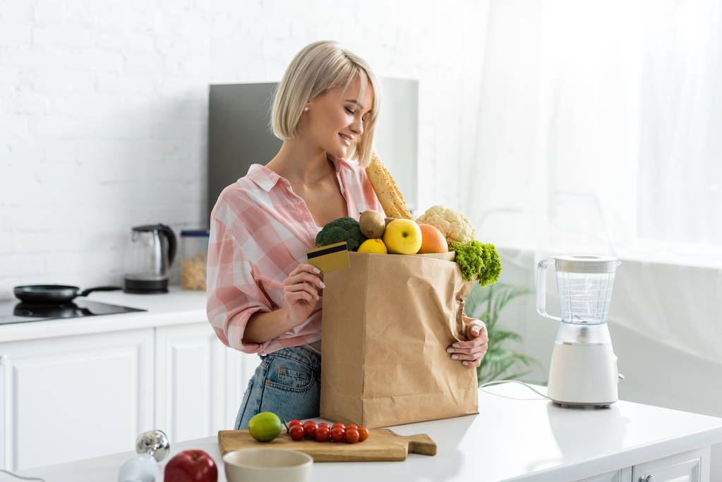 glückliche blonde junge Frau mit Kreditkarte in der Nähe einer Papiertüte mit Lebensmitteln  - Foto, Bild