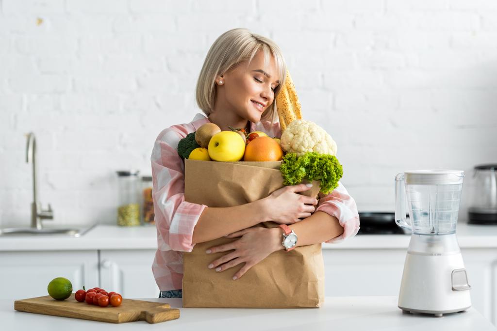 glückliche blonde junge Frau umarmt Papiertüte mit Lebensmitteln  - Foto, Bild