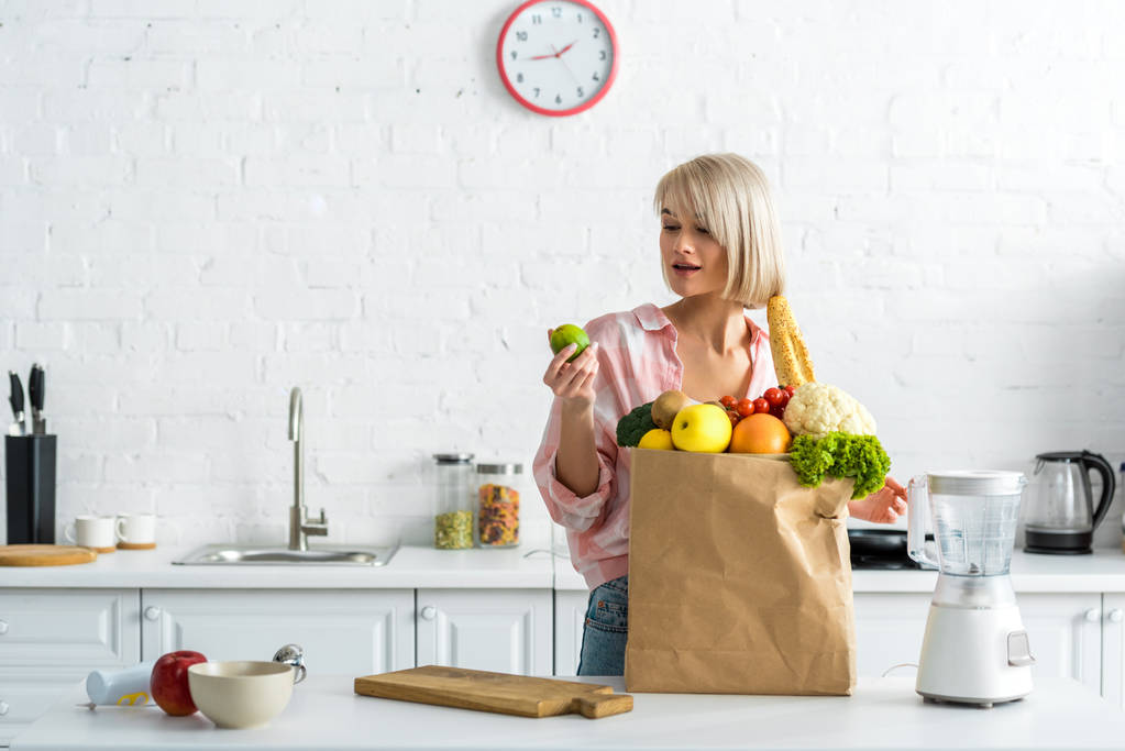 mujer rubia sorprendida sosteniendo cal cerca de bolsa de papel con comestibles
  - Foto, Imagen