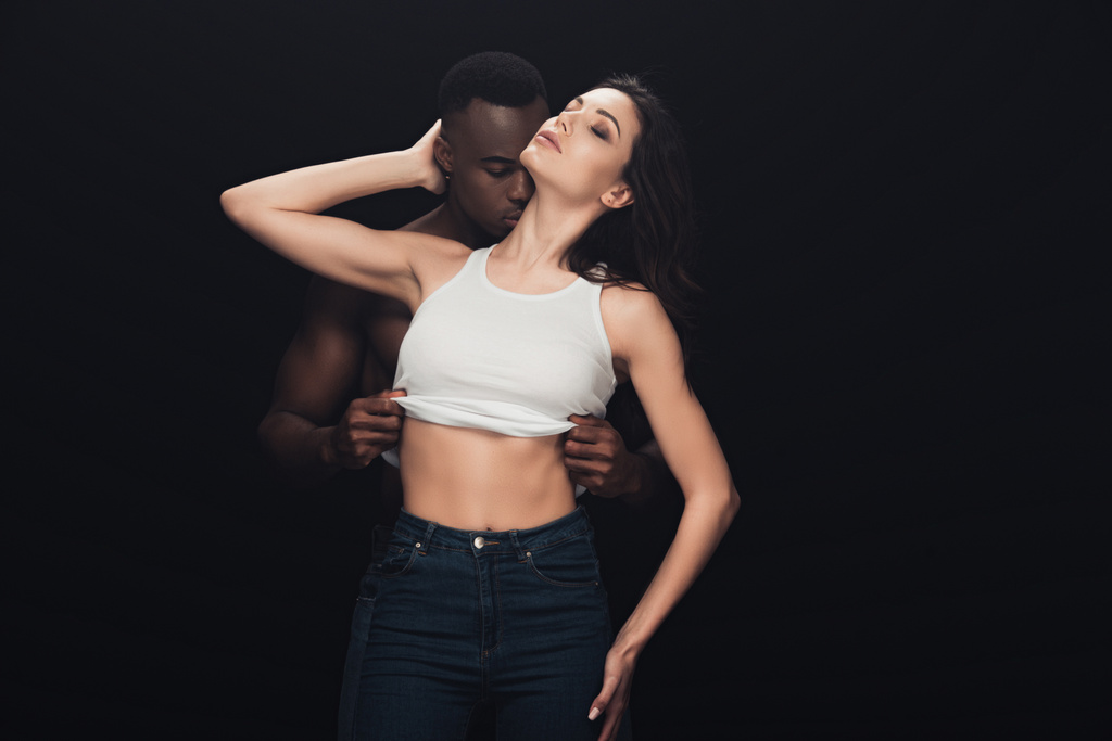 knappe Afro-Amerikaanse man uitkleden mooie sensuele vrouw geïsoleerd op zwart met kopieerruimte - Foto, afbeelding