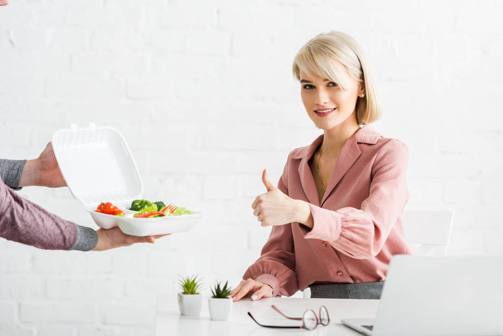 bijgesneden weergave van de mens Holding afhaalmaaltijden doos met groenten in de buurt van Happy blonde meisje toont thumb up - Foto, afbeelding