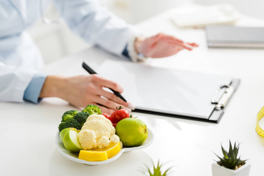 abgeschnittene Ansicht eines Ernährungswissenschaftlers, der Stift in der Nähe von Klemmbrett und Teller mit Bio-Lebensmitteln hält  - Foto, Bild