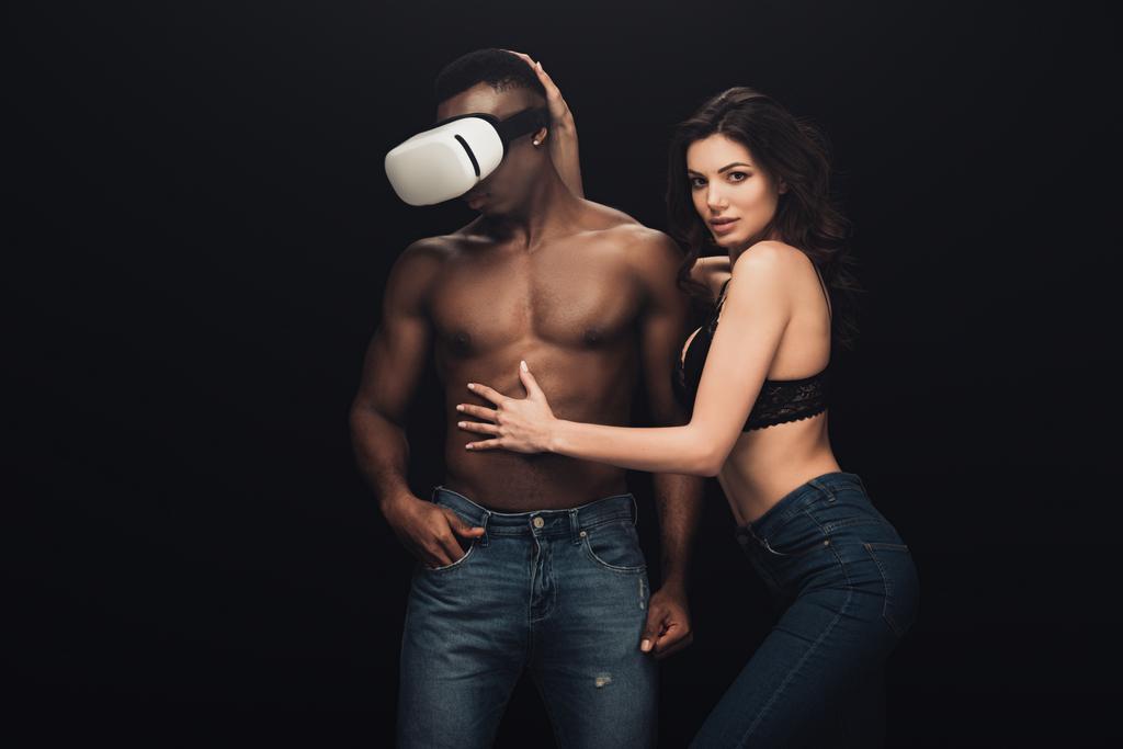 schöne sexy Frau posiert und schaut in die Kamera in der Nähe afrikanischer amerikanischer Mann in Virtual-Reality-Headset isoliert auf schwarz - Foto, Bild