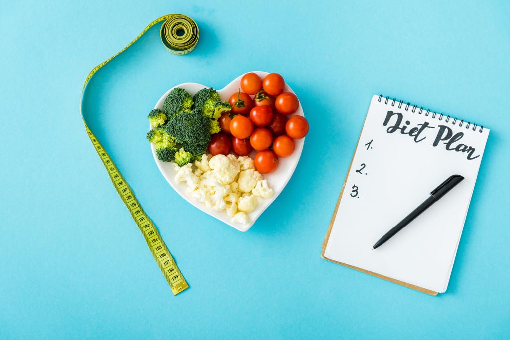 vista dall'alto di verdure saporite su piatto a forma di cuore vicino a nastro di misurazione e notebook con iscrizione di piano di dieta su azzurro
 - Foto, immagini