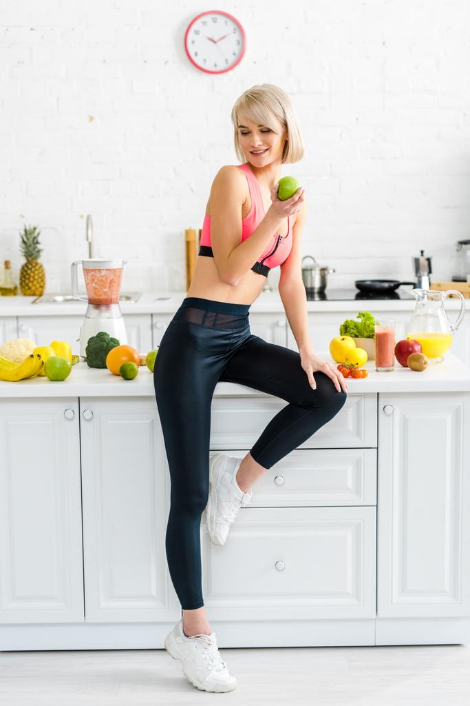 attraktive Frau in Sportkleidung mit Apfel in der Nähe von Zutaten in der Küche  - Foto, Bild