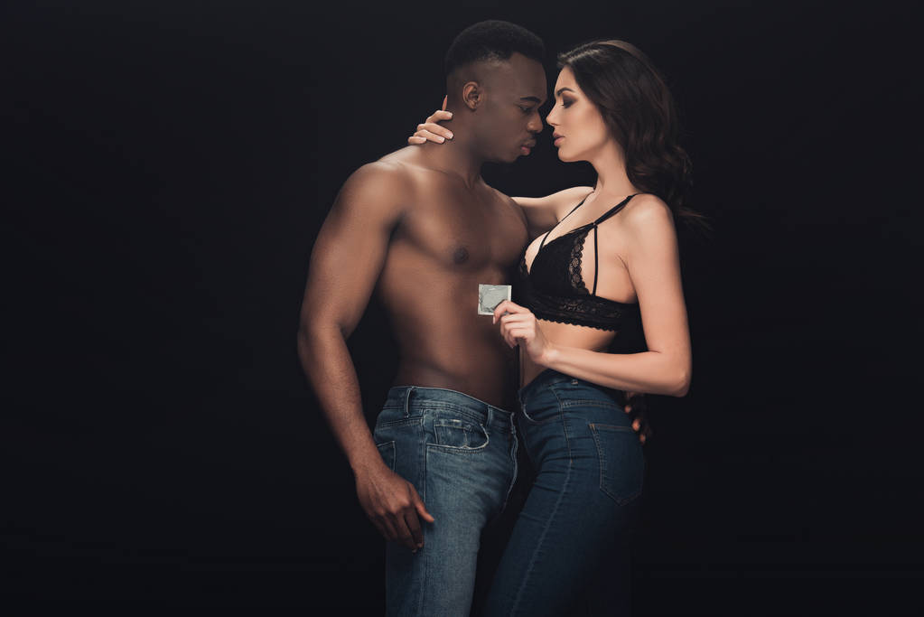mooie sexy vrouw omarmen hemdloze Afro-Amerikaanse man en holding condoom geïsoleerd op zwart - Foto, afbeelding