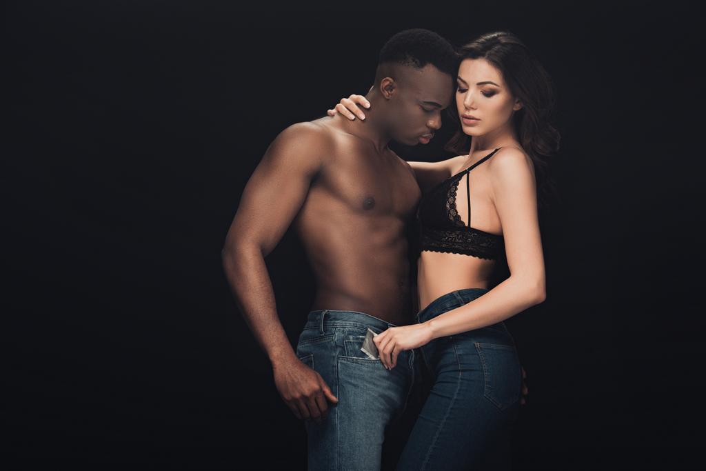 魅力的な女性が上半身裸アフリカ系アメリカ人の男性を抱きしめ、コンドームを黒に分離した - 写真・画像