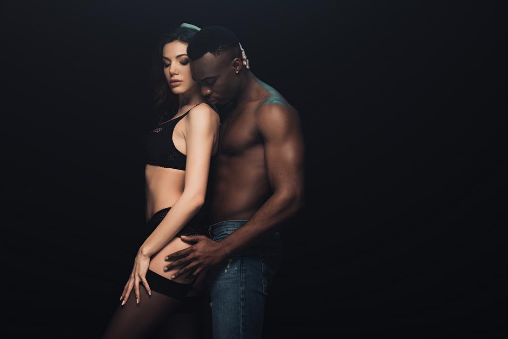 african american man hugging beautiful sexy woman in lingerie isolated on black with copy space - Photo, Image