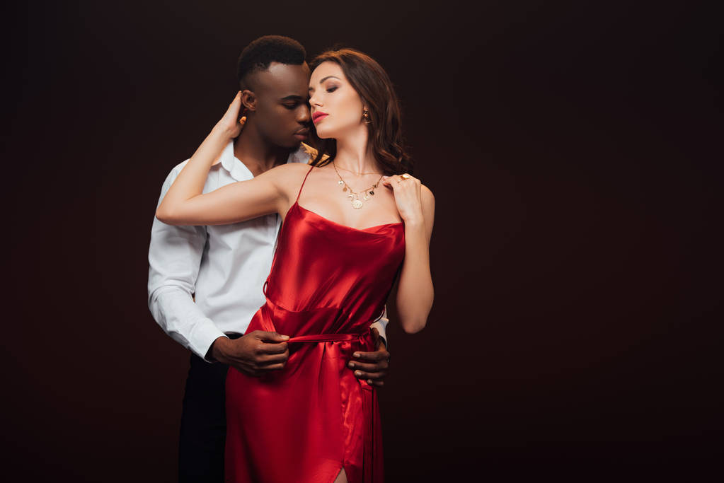 hombre afroamericano abrazando hermosa mujer seductora en vestido rojo aislado en negro con espacio de copia
 - Foto, Imagen