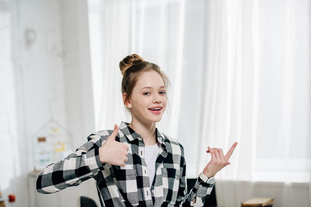 Uśmiechnięta Nastolatka w Checkered shirt pokazując kciuki w górę w domu - Zdjęcie, obraz