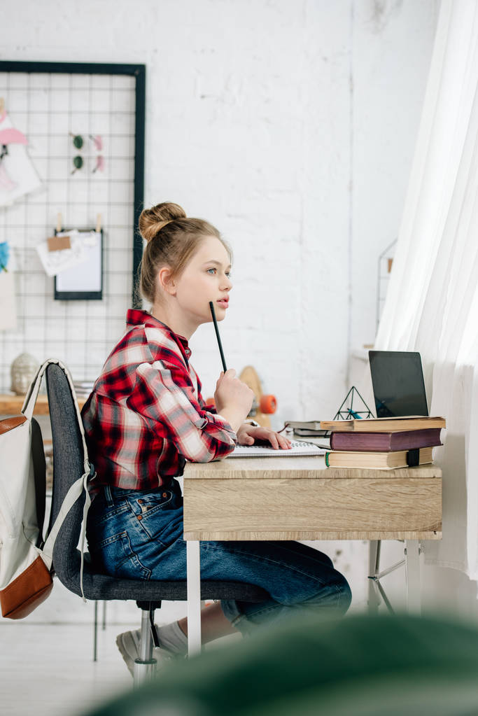 Pensive teenager in checkered shirt holding pen at table while doing homework - Photo, Image