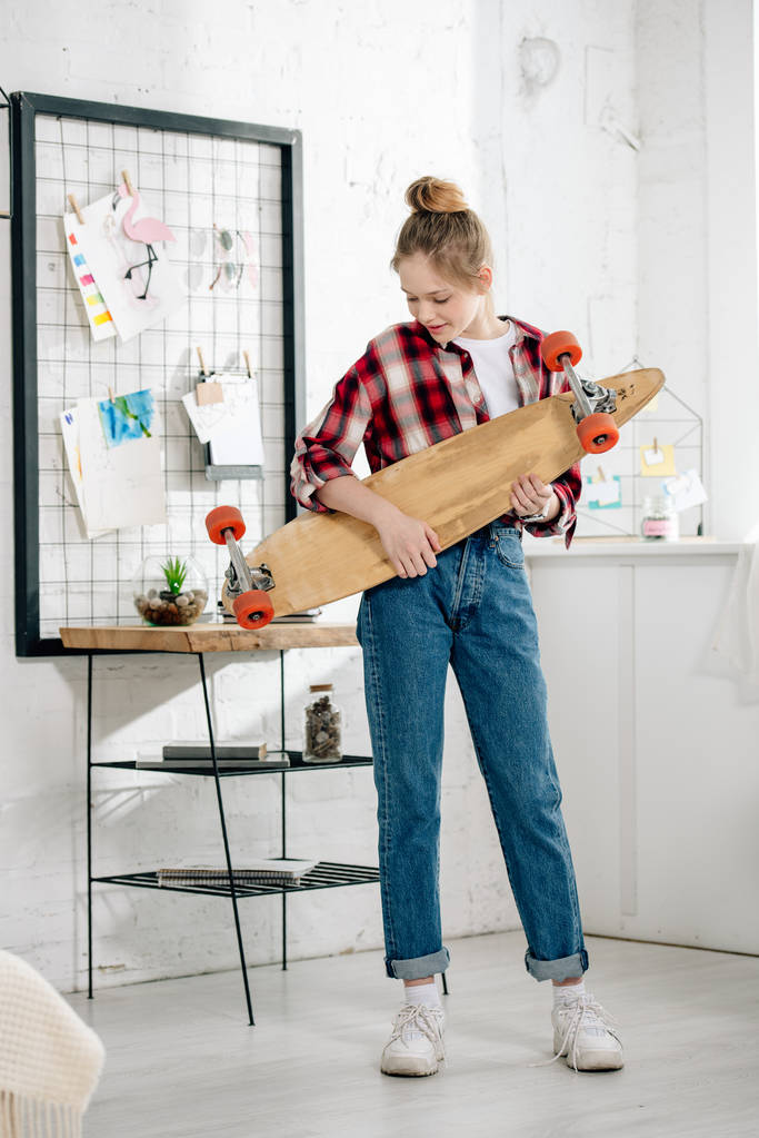 Vue complète de l'adolescent en jeans tenant longboard à la maison
 - Photo, image