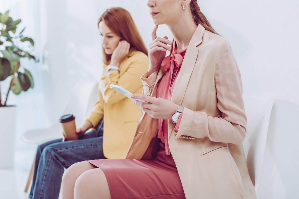 обрезанный вид задумчивой женщины с помощью смартфона рядом с сотрудником с бумажной чашкой
  - Фото, изображение