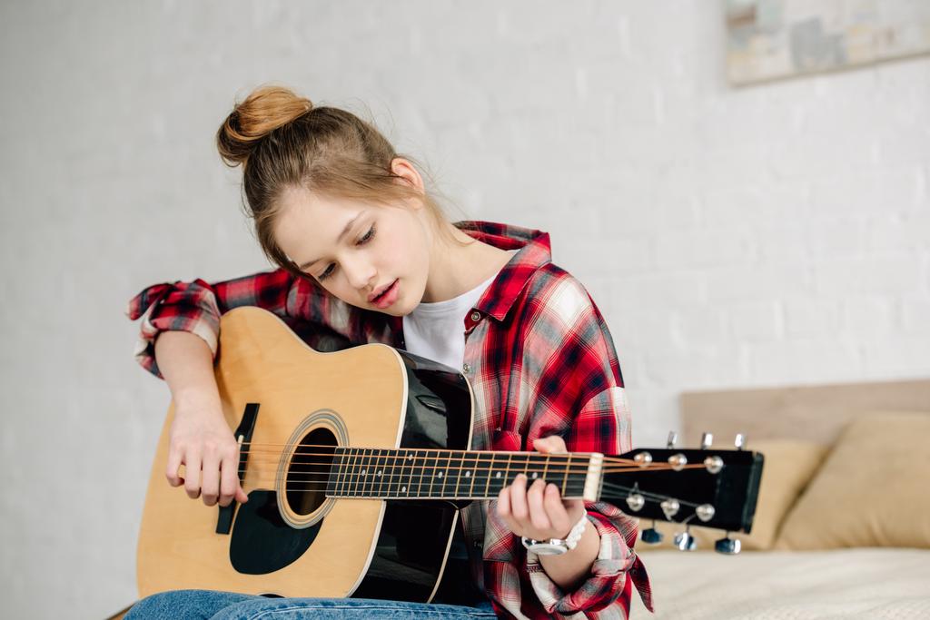 Adolescente concentrado en camisa a cuadros tocando la guitarra acústica en casa
 - Foto, Imagen