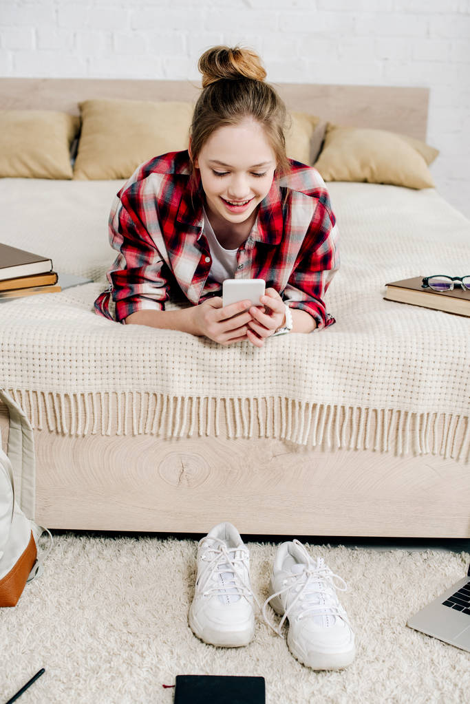 Adolescente sonriente con camisa a cuadros acostada en la cama y usando un teléfono inteligente
 - Foto, Imagen