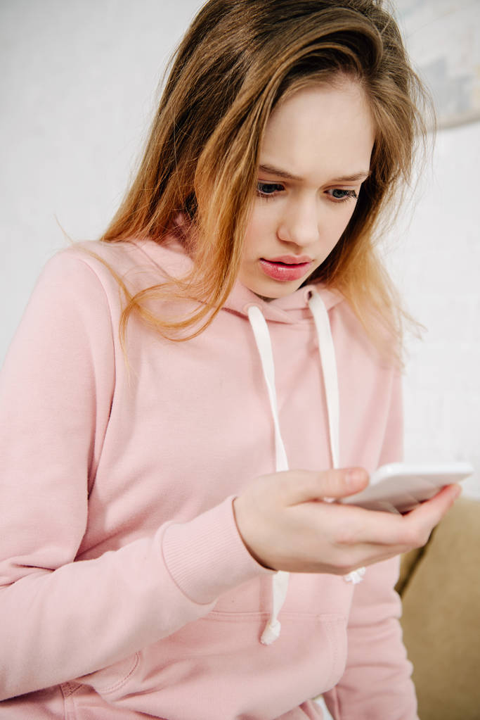 Meghökkent tizenéves gyerek rózsaszín kapucnis segítségével smartphone otthon - Fotó, kép
