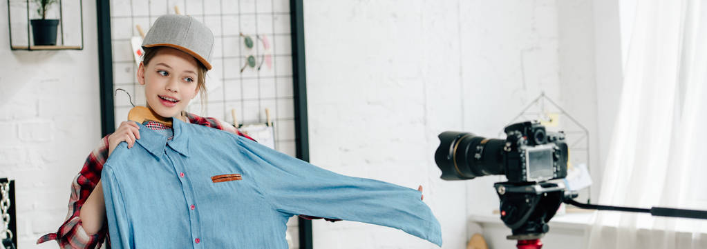 Panoramatický záběr veselého teenagera v čepici zobrazující košili u videokamery - Fotografie, Obrázek