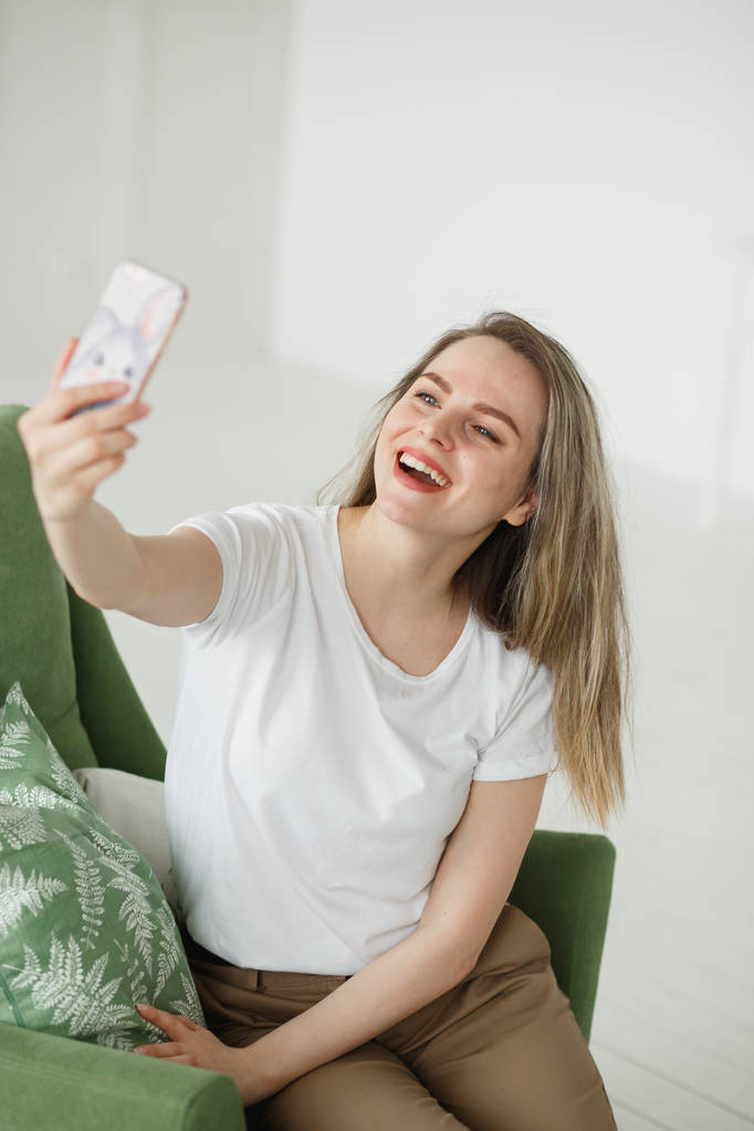 Πορτρέτο του όμορφη νεαρή γυναίκα λήψη selfie στην πολυθρόνα στο ανοιχτό σαλόνι. - Φωτογραφία, εικόνα