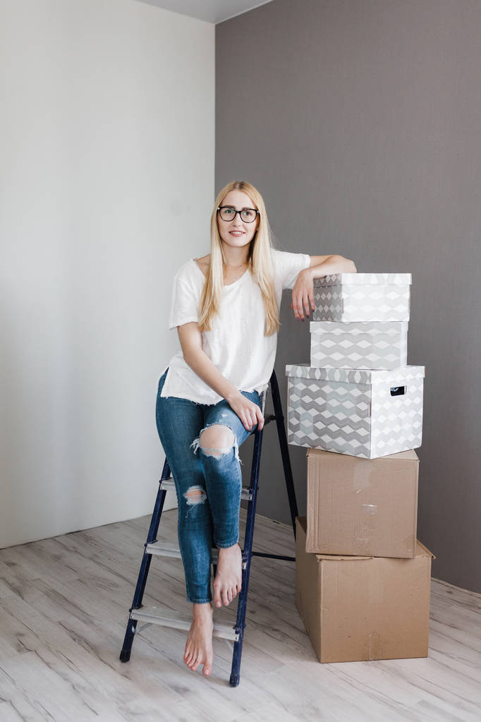 Jeune femme emménageant dans sa nouvelle maison et faisant une cure de jouvence à la maison, elle est assise sur l'échelle avec de nombreuses boîtes en carton autour
 - Photo, image