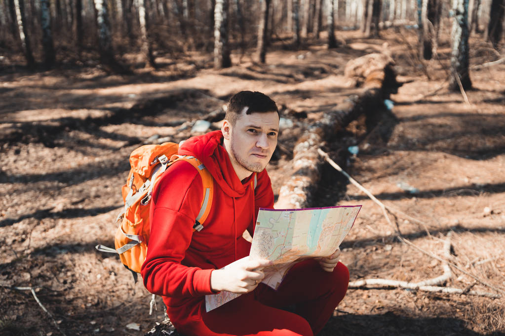 Άνδρας με ανάγνωση χάρτη σε woodsyoung ενέπνευσε ο άνθρωπος στο κόκκινο sportswear κάθεται σε πεσμένο δέντρο κορμών στο δάσος και ανάγνωση χάρτη ενώ ταξιδεύουν με το σακίδιο σακίδιο - Φωτογραφία, εικόνα