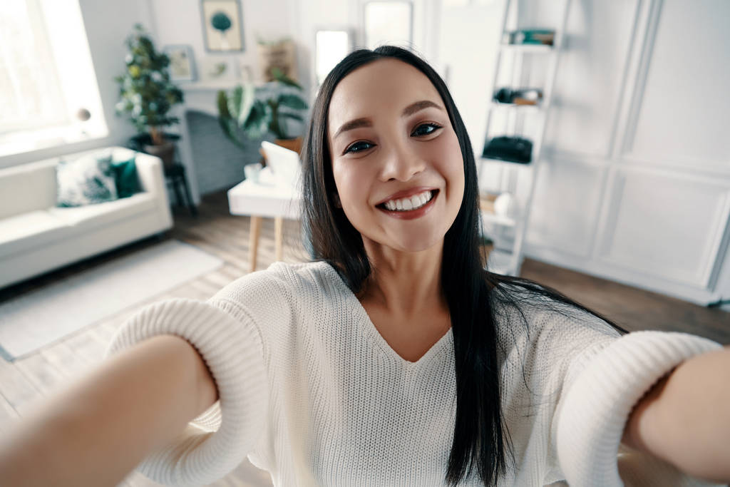 Perfekcyjny selfie. Atrakcyjna młoda kobieta patrząc na kamerę i uśmiechając się podczas robienia selfie w domu             - Zdjęcie, obraz