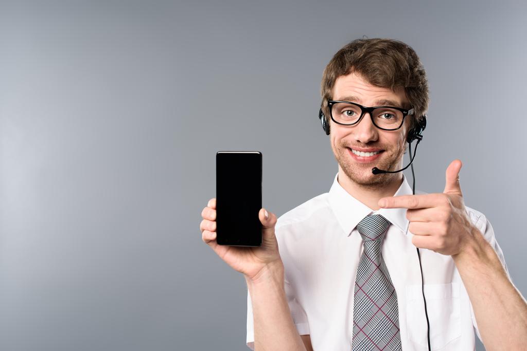 operador del centro de llamadas sonriente en gafas y auriculares apuntando con el dedo al teléfono inteligente con pantalla en blanco
 - Foto, Imagen