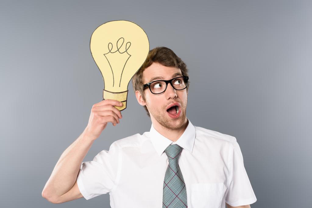 zasněný podnikatel v brýlích drží žlutý papír vyjmout žárovku na šedém pozadí - Fotografie, Obrázek