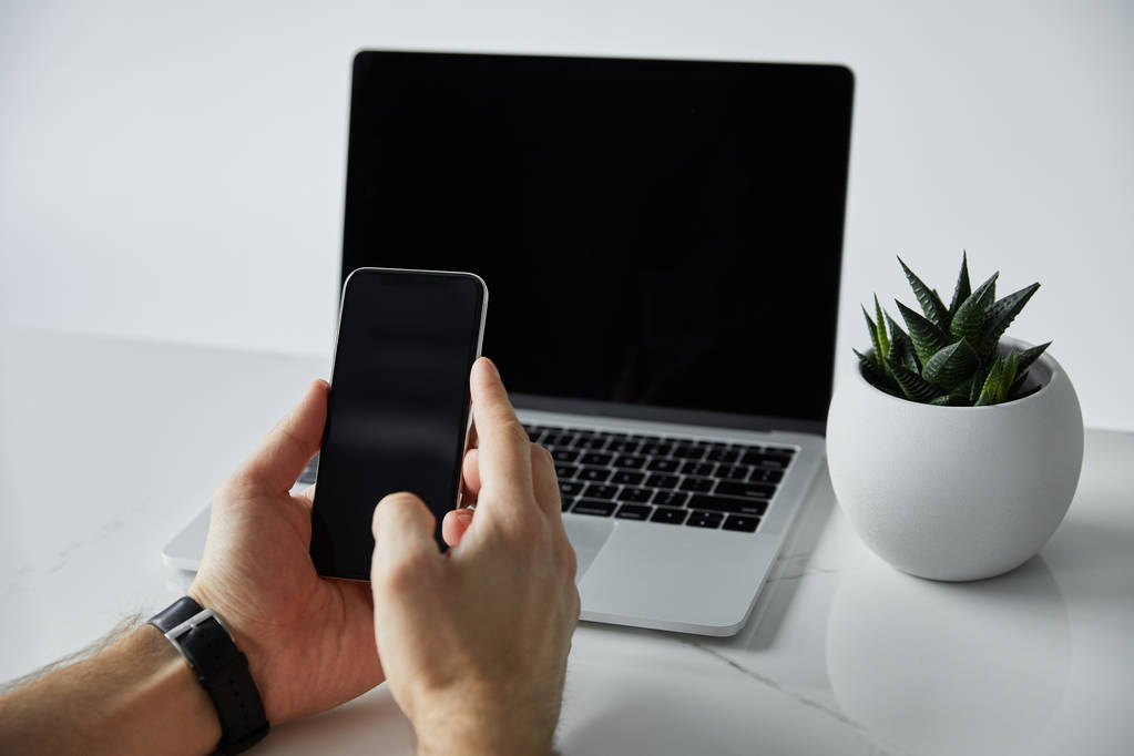 частковий вид людини, що використовує смартфон біля ноутбука з порожнім екраном, і горщик для квітів ізольований на сірому
 - Фото, зображення