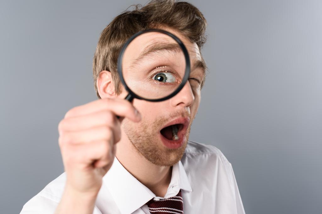 geschokt zakenman met grappige gezicht expressie op zoek in Vergrootglas op grijze achtergrond - Foto, afbeelding