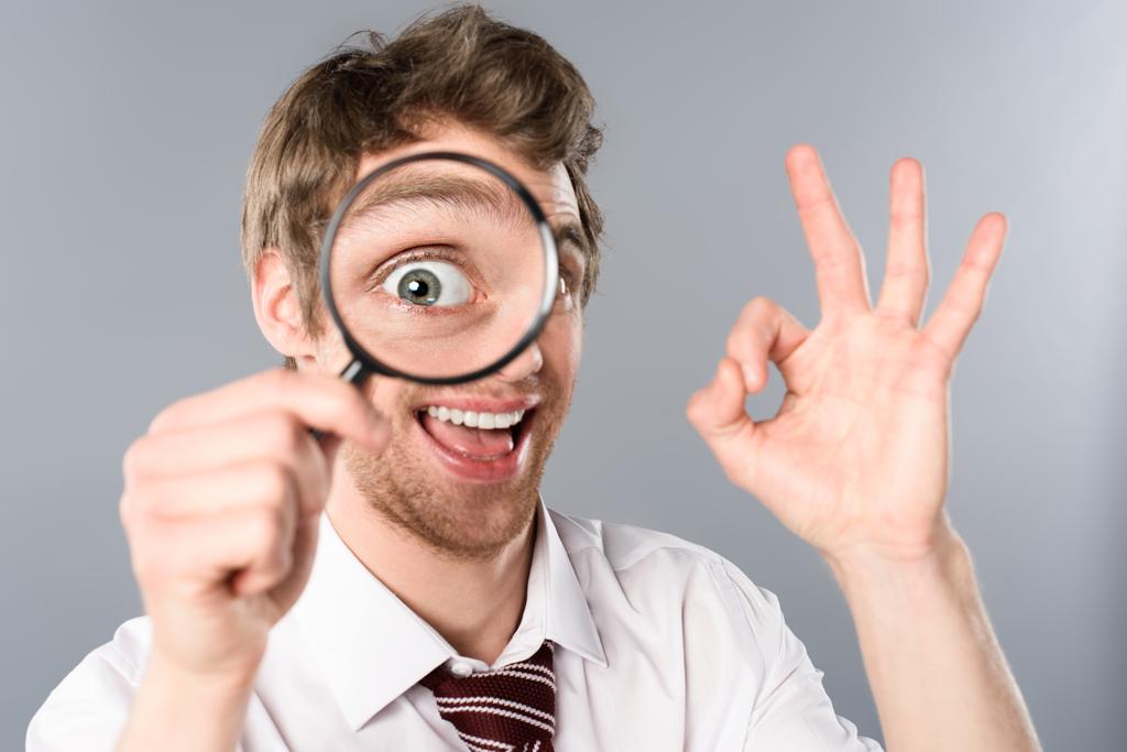 positive businessman with funny face expression looking in magnifier and showing ok sign on grey background - Photo, Image