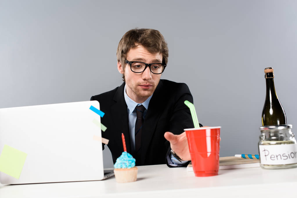 усталый бизнесмен, сидящий на рабочем месте с кексом и глядя на бумажную чашку изолированы на серый
 - Фото, изображение