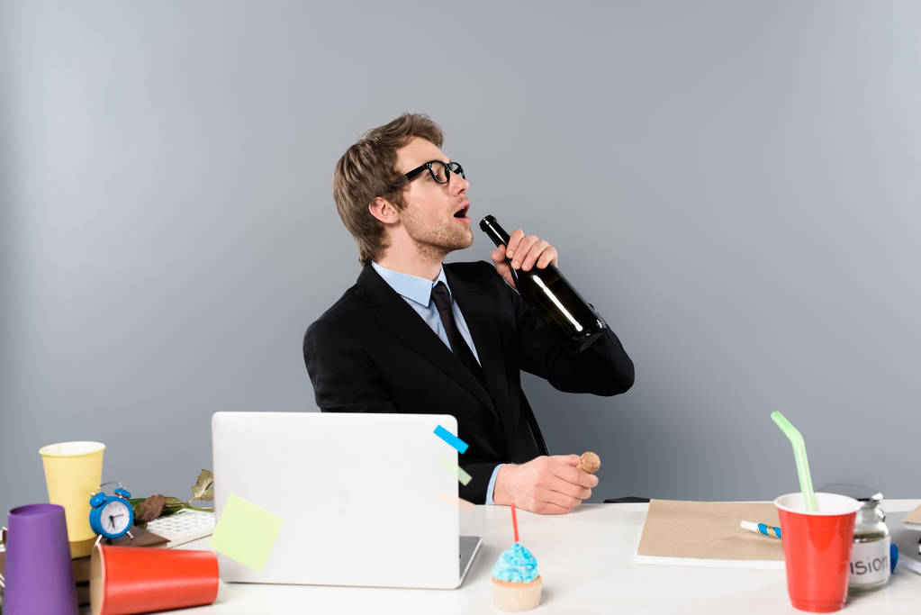 бизнесмен, сидящий на рабочем месте с кексом и пьющий шампанское из бутылки изолированы на сером
 - Фото, изображение