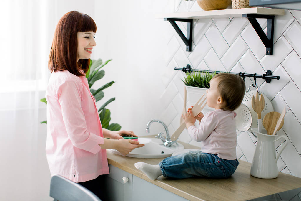 Young happy mother and her cute curly toddler daughter having fun together in a white kitchen. Mother is washing the dishes, plates, while baby girl is sitting near sink and playing with wooden fork - Photo, Image