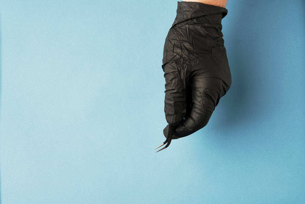 Χέρι-μαύρο γάντι εκμετάλλευση τσιμπιδάκια σε ανοιχτό μπλε φόντο - Φωτογραφία, εικόνα