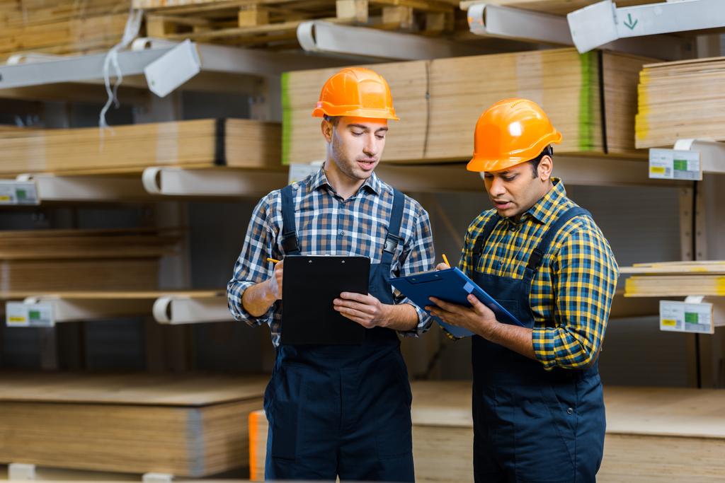 two multicultural warehouse workers in uniform writing in clipboards  - Photo, Image