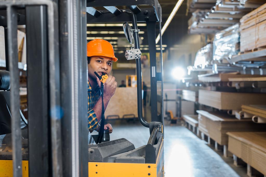 przystojny indyjski pracownik siedzący w wózek widłowy maszyna w Warehouse i rozmowa na walkie talkie - Zdjęcie, obraz