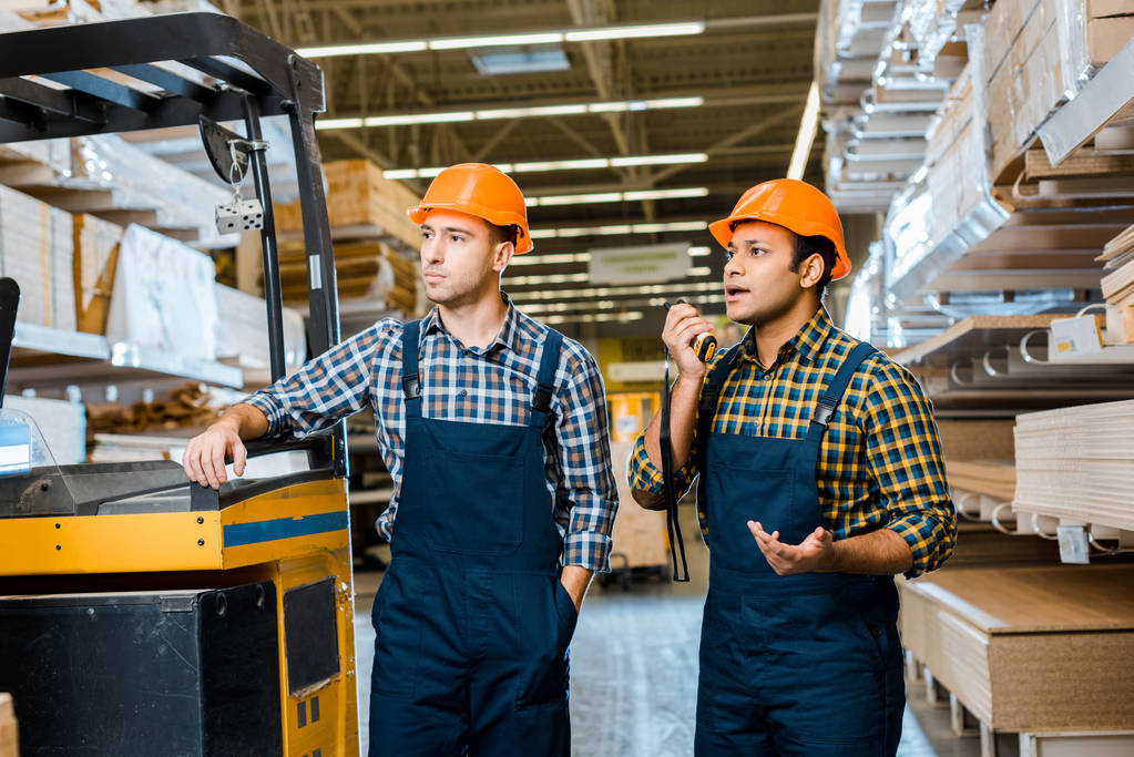 multiculturele Warehouse Workers in uniform en helmen staande in de buurt van vorkheftruck machine in pakhuis - Foto, afbeelding