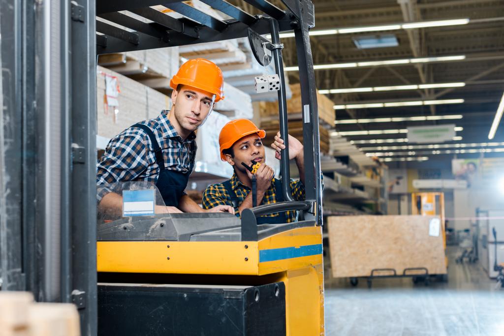 attentive worker sitting in forklift machine near his indian colleague talking on walkie talkie - Photo, Image
