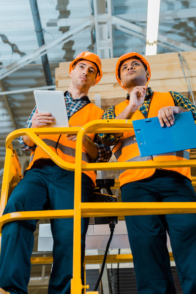 concentrated multicultural warehouse workers with digital tablet and clipboard standing on scissor lift  - Photo, Image